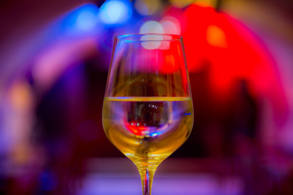 Auf dem Bild ist ein mit Weißwein gefülltes Weinglas vor buntem Scheinwerferlicht zu sehen.