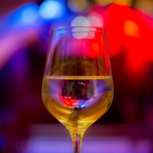 Auf dem Bild ist ein mit Weißwein gefülltes Weinglas vor buntem Scheinwerferlicht zu sehen.
