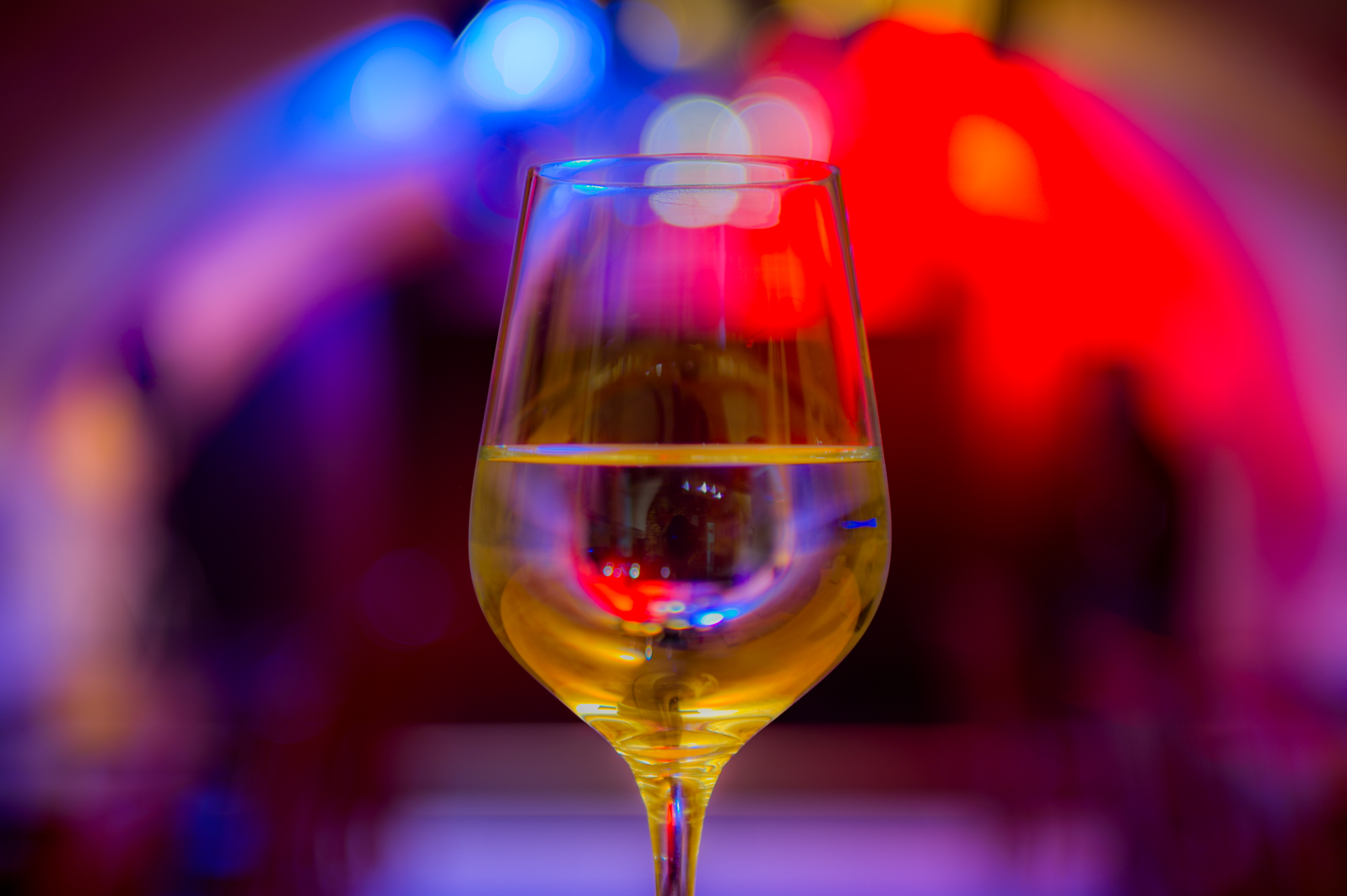 Auf dem Bild ist ein mit Weißwein gefülltes Weinglas vor buntem Scheinwerferlicht zu sehen.