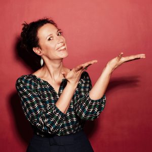 Auf dem Foto ist die Musikkabarettistin Alice Köfer vor rotem Hintergrund zu sehen.