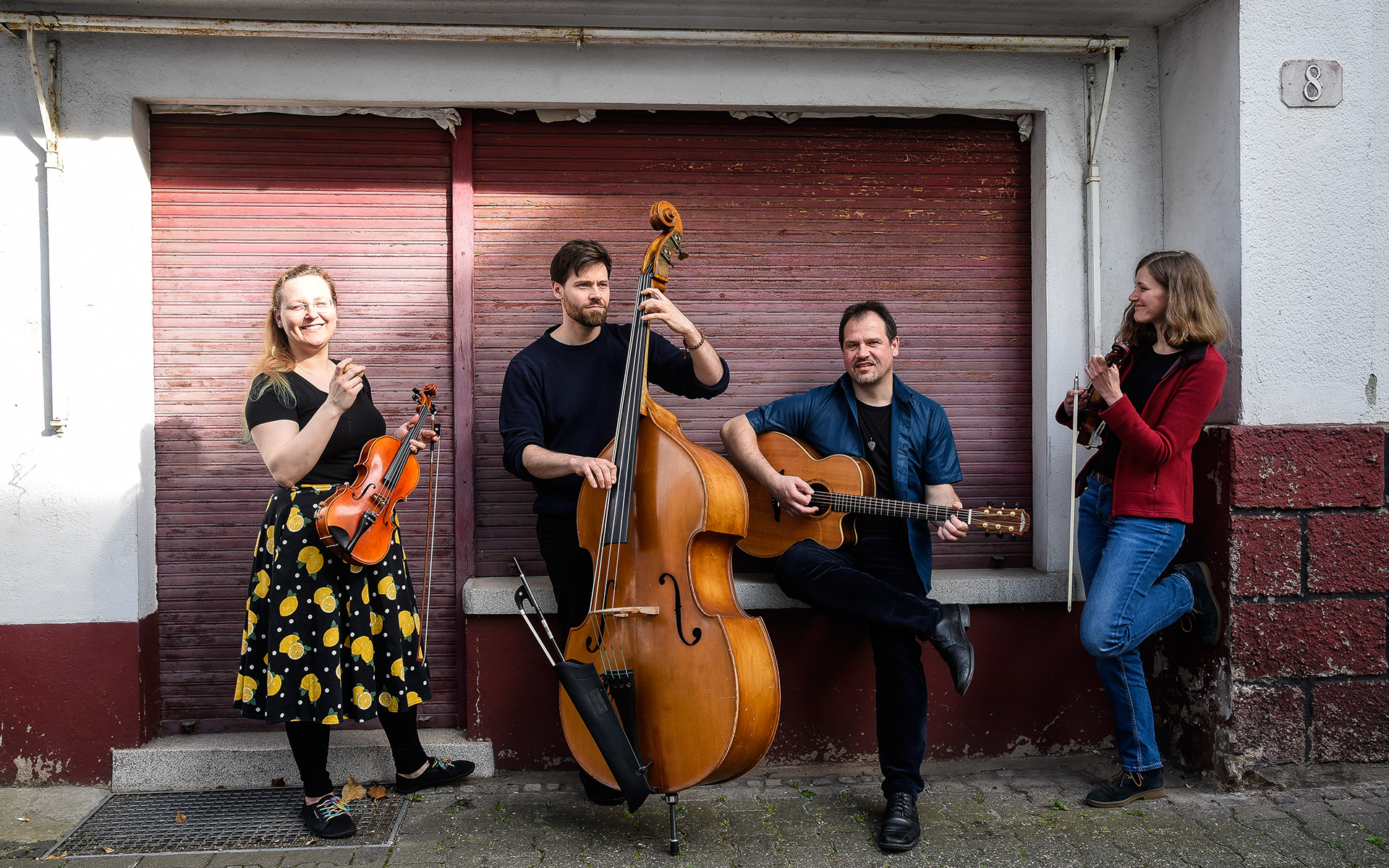 Auf dem Bild ist das Quartett Feinschmeckerfolk mit seinen Instrumenten vor einem alten heruntergelassenen Rolladen zu sehen.