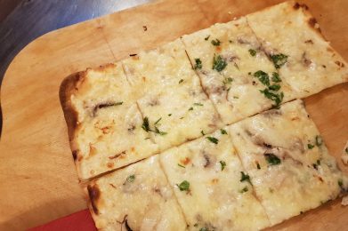 Auf dem Bild ist ein Flammkuchen auf dem Holzbrett zu sehen.