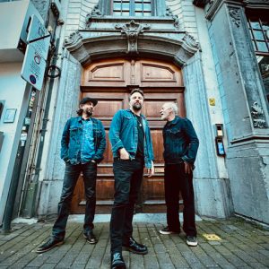 Auf dem Bild sind die drei Musiker vom Gerry Hundt Trio vor einem mächtigen Holzportal stehend zu sehen.