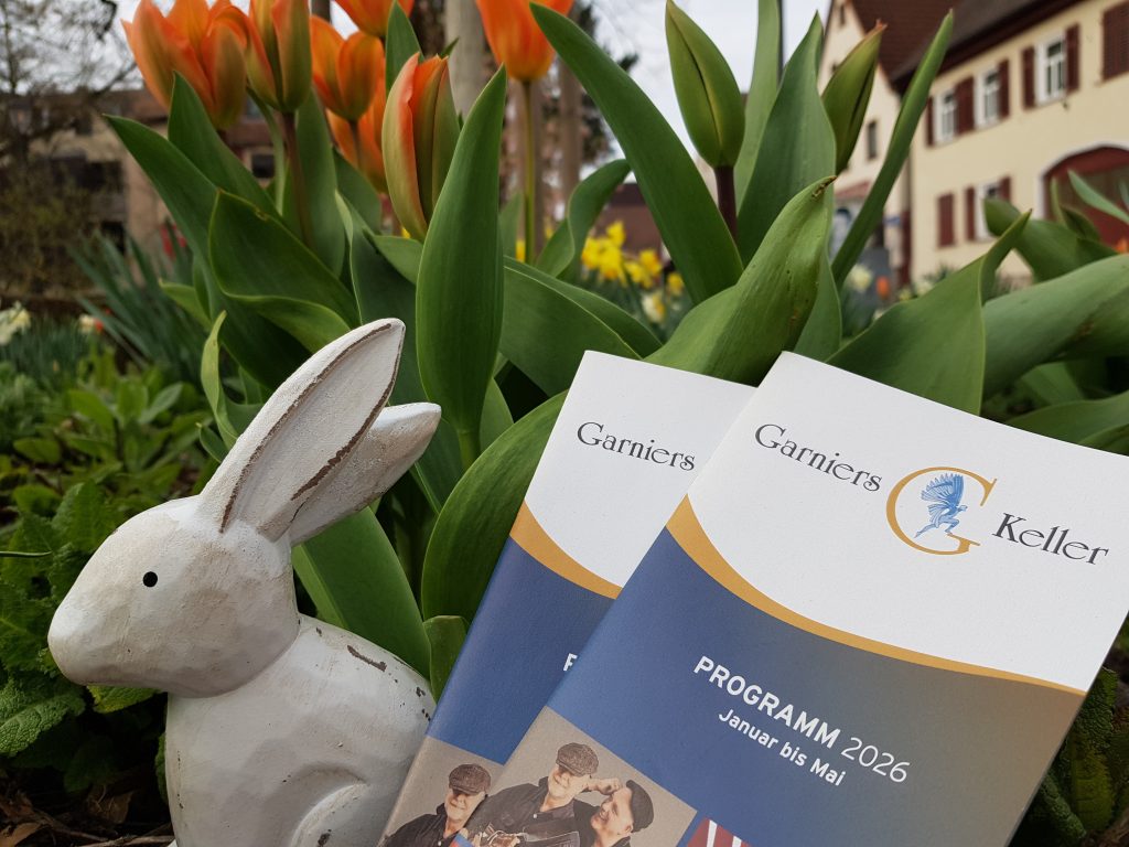 Auf dem Foto sind ein Holzhase und Programmhefte vom Garniers Keller vor Tulpen zu sehen.