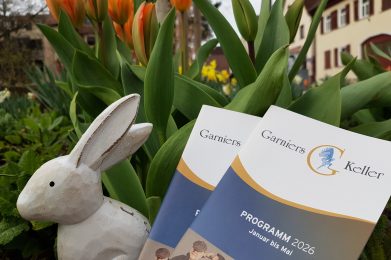 Auf dem Foto sind ein Holzhase und Programmhefte vom Garniers Keller vor Tulpen zu sehen.