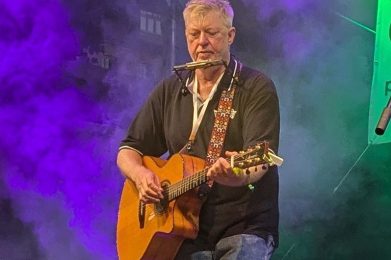 Auf dem Bild ist Paddy Schmidt mit Gitarre und Mundharmonika zu sehen.