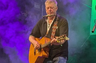 Auf dem Bild ist Paddy Schmidt mit Gitarre und Mundharmonika zu sehen.