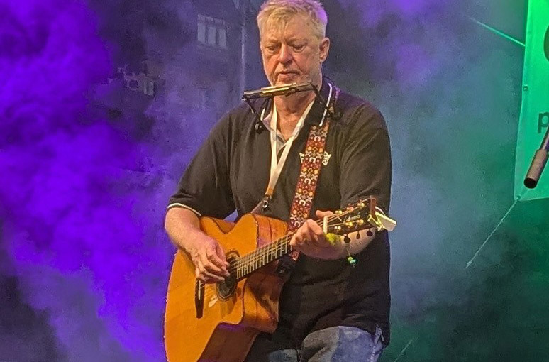 Auf dem Bild ist Paddy Schmidt mit Gitarre und Mundharmonika zu sehen.