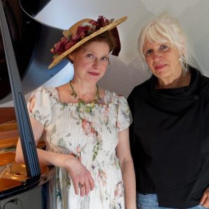 Auf dem Bild sind Jenny Rehs in Blumenkleid und Hut und Waltraut Bartl vor einem Flügel stehend zu sehen.