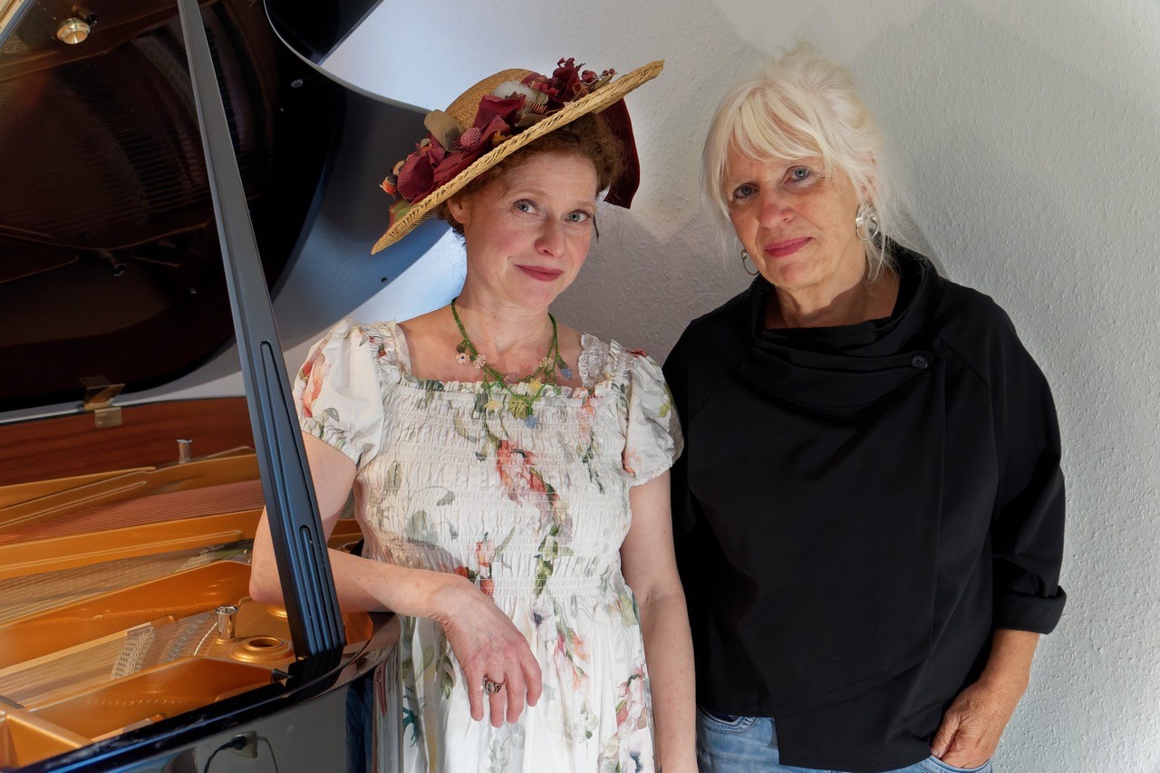 Auf dem Bild sind Jenny Rehs in Blumenkleid und Hut und Waltraut Bartl vor einem Flügel stehend zu sehen.