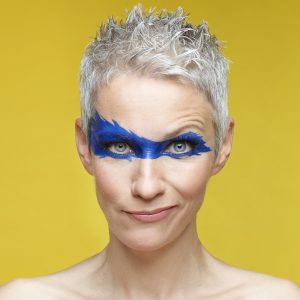 Auf dem Bild ist Tina Häussermann vor gelbem Hintergrund mit blau aufgemalter Gesichtsmaske zu sehen.