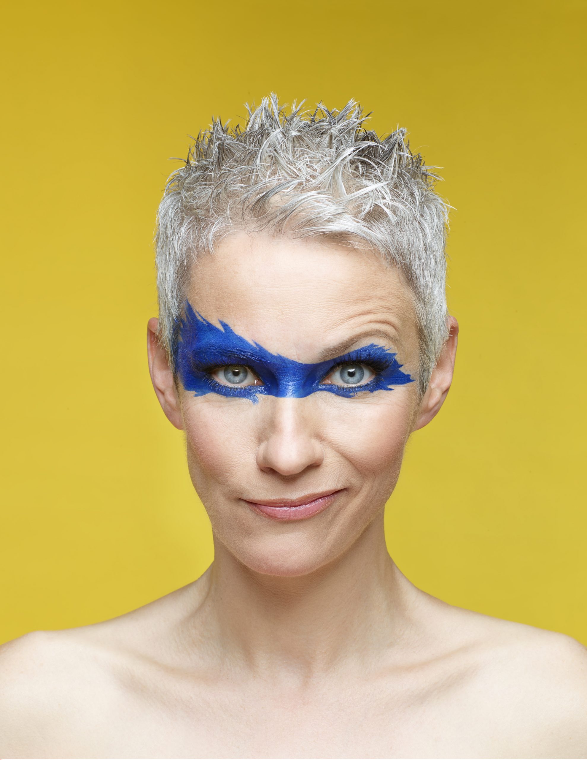 Auf dem Bild ist Tina Häussermann vor gelbem Hintergrund mit blau aufgemalter Gesichtsmaske zu sehen.